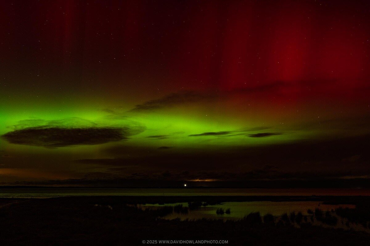 The aurora borealis illuminates the sky in bright green and red over the lit beacon of Sandy Neck Lighthouse.
