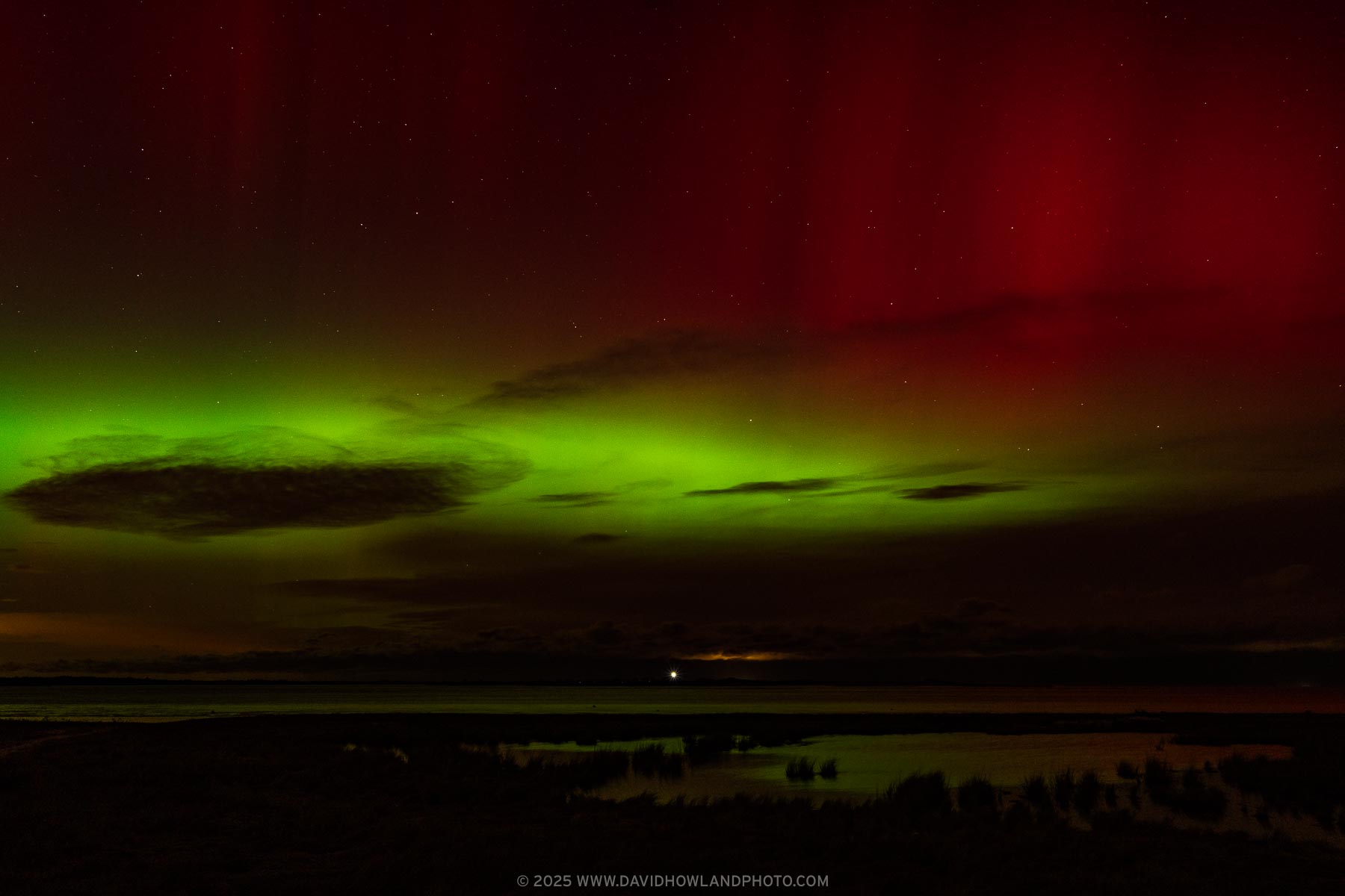 The aurora borealis illuminates the sky in bright green and red over the lit beacon of Sandy Neck Lighthouse.