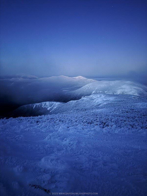 A twilight view from a snow-covered summit looking across Franconia Notch toward the Kinsman Range in the White Mountains, with the landscape bathed in deep blue tones, fog drifting through the valleys, and faint stars emerging in the darkening sky.
