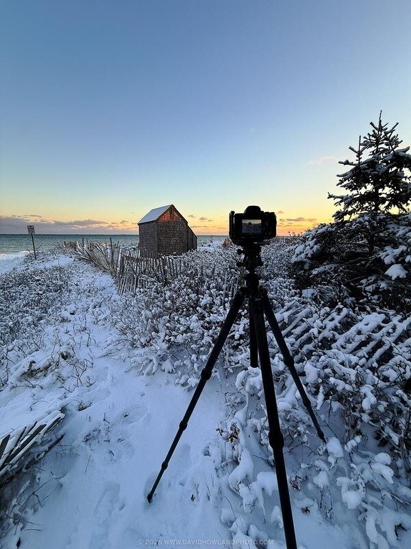 A camera on a tripod stands in a snow-covered beach path at Forest Beach in Chatham, MA, aimed at the weathered wooden beach shack and driftwood fence near the ocean under a soft orange and blue winter sunset sky.