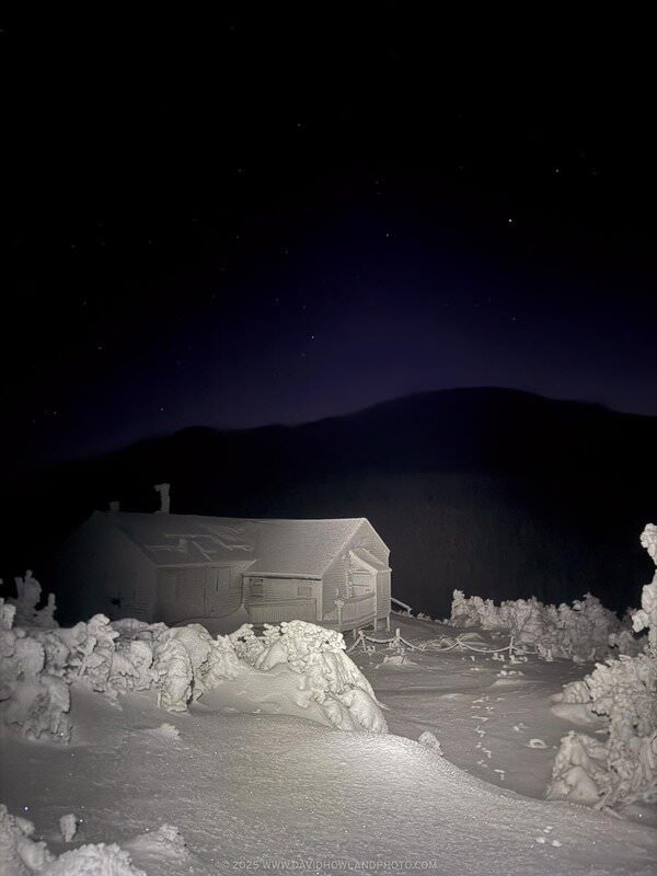A snow-covered Greenleaf Hut sits on a winter landscape at night, surrounded by frost-laden evergreen trees, with a dark mountain silhouette and starry sky in the background.