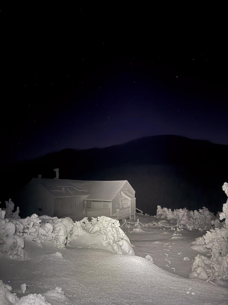 A snow-covered Greenleaf Hut sits on a winter landscape at night, surrounded by frost-laden evergreen trees, with a dark mountain silhouette and starry sky in the background.