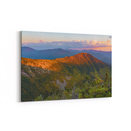 A mountain landscape at sunset showing Bondcliff peak illuminated in golden-orange light in the foreground, with layers of blue-purple mountain ridges receding into the distance beneath a pink and orange sky with scattered clouds.