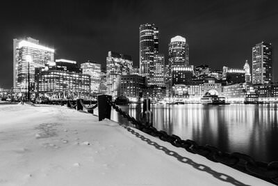 A black and white photo of the illuminated Boston skyline above the waters of Boston Harbor and a snow covered walkway.