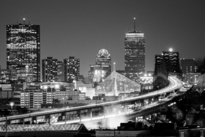 A black and white photo of the illuminated skyscrapers and bridges of Boston, Massachusetts at night.