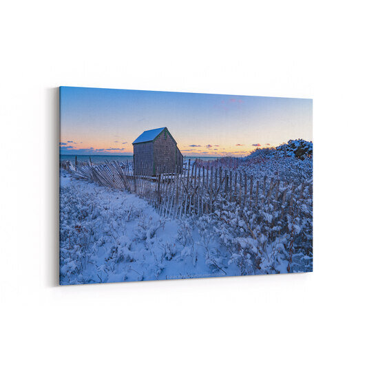 A weathered shingled shed stands amid snow-covered dunes and wooden snow fencing on a Cape Cod beach in Chatham, Massachusetts, with the ocean and a pastel winter sunset visible in the background.