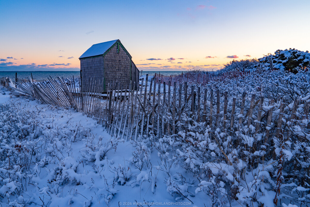 A weathered shingled shed stands amid snow-covered dunes and wooden snow fencing on a Cape Cod beach in Chatham, Massachusetts, with the ocean and a pastel winter sunset visible in the background.