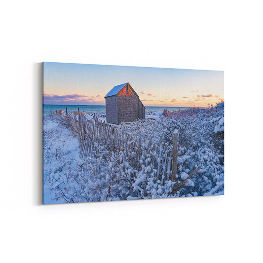 A weathered gray shingled shed stands on a snow-covered beach at sunset, surrounded by wooden snow fences and frost-laden beach grasses, with the ocean and pastel purple-and-orange sky visible in the background.
