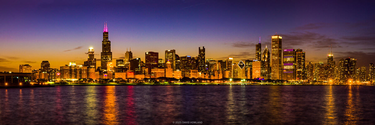 A panorama photo of the skyscrapers of the Chicago city skyline lit up against the fading orange sunset light.