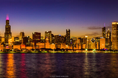 A panorama photo of the skyscrapers of the Chicago city skyline lit up against the fading orange sunset light.