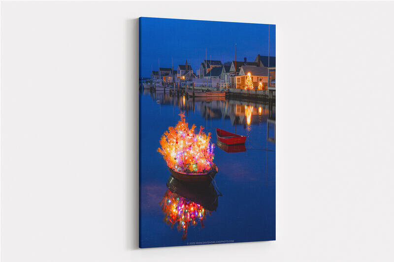 A small dinghy decorated with a multicolored illuminated Christmas tree topped with a star floats in calm blue harbor water at twilight, with a red dinghy and waterfront homes with holiday lights reflected in the background.