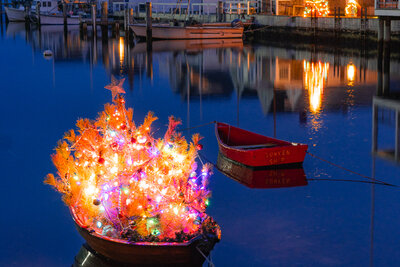 A small dinghy decorated with a multicolored illuminated Christmas tree topped with a star floats in calm blue harbor water at twilight, with a red dinghy and waterfront homes with holiday lights reflected in the background.