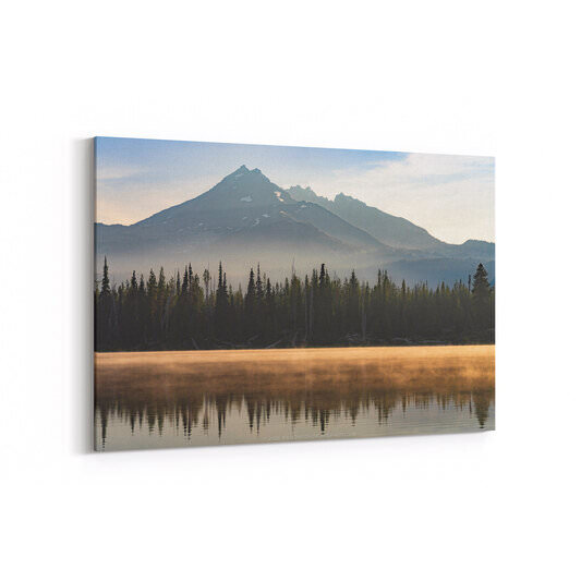 A view across Sparks Lake at dawn showing Broken Top, a jagged mountain peak with patches of snow, in the background; a line of dense pine forest along the near shoreline; and a band of amber-tinted mist hovering just above the lake's calm surface, with the mountain and trees reflected in the water below it.