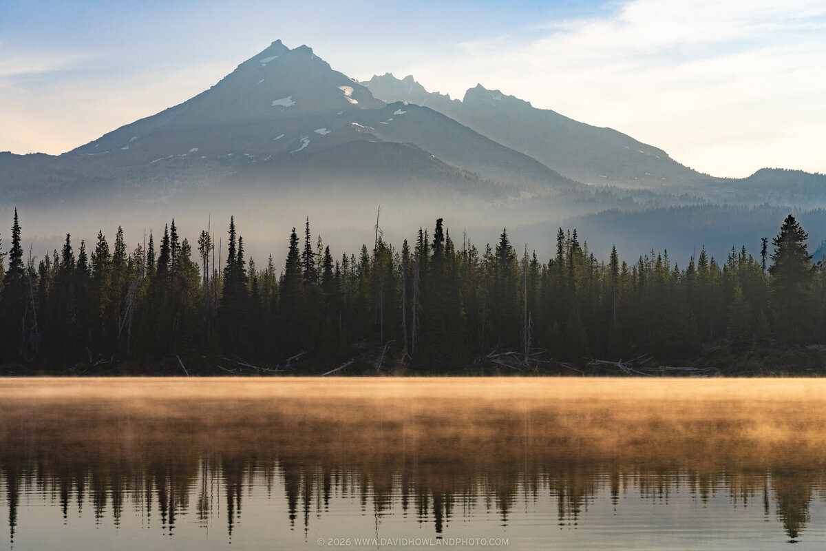 A view across Sparks Lake at dawn showing Broken Top, a jagged mountain peak with patches of snow, in the background; a line of dense pine forest along the near shoreline; and a band of amber-tinted mist hovering just above the lake's calm surface, with the mountain and trees reflected in the water below it.