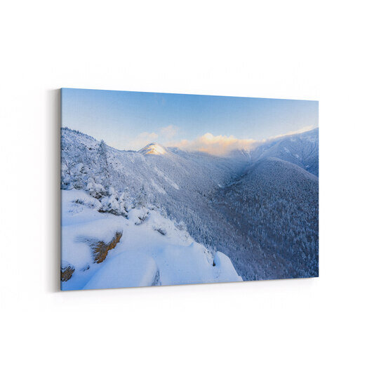 A winter mountain view at dawn shows snow-covered peaks and forested slopes under a pale blue sky, with warm golden sunlight illuminating one distant summit while the valley below remains in shadow.