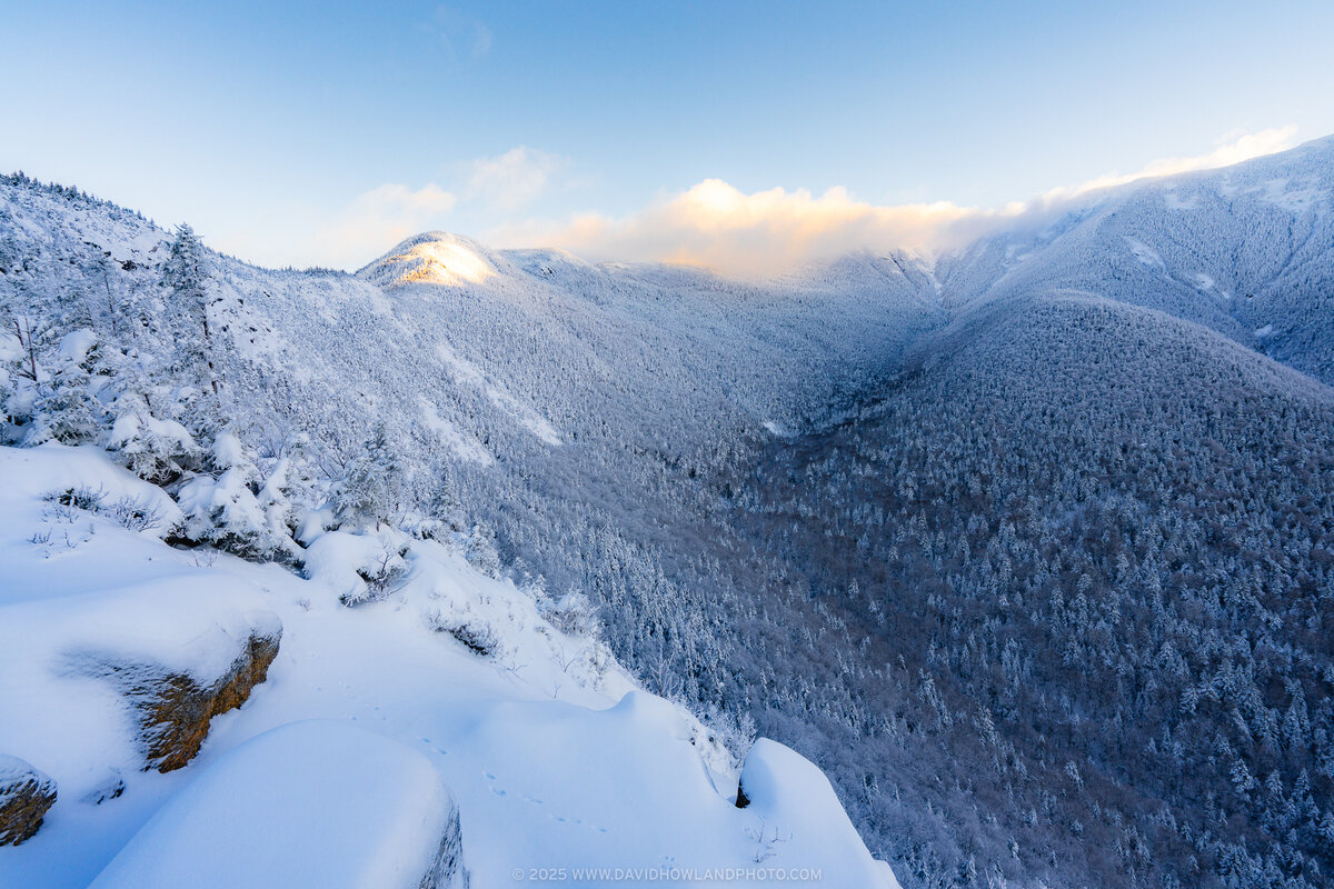 A winter mountain view at dawn shows snow-covered peaks and forested slopes under a pale blue sky, with warm golden sunlight illuminating one distant summit while the valley below remains in shadow.