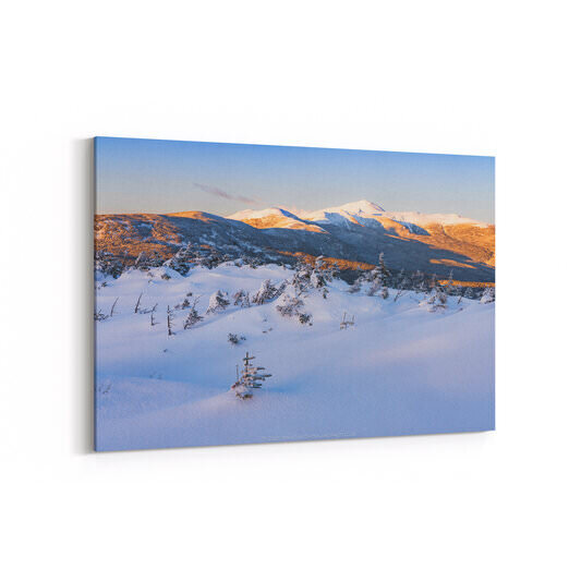 A sweeping dusk view of the Southern Presidential Range, with a vast snow-covered alpine foreground dotted with small frost-covered conifers giving way to forested slopes bathed in warm golden light, and Mount Washington's snow-capped summit rising prominently against a clear pale blue sky in the distance, with Mounts Pierce, Eisenhower, and Monroe also visible along the ridge.