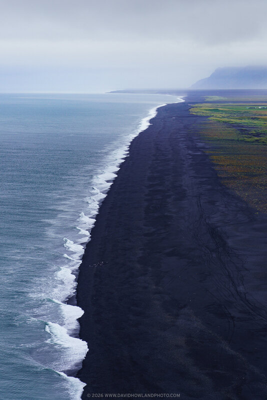 A sweeping view shows waves rolling in along Iceland’s Dyrhólaey Black Sand Beach, where a long, dark shoreline curves into the distance beside gray-blue ocean water, green coastal fields, and misty cliffs under an overcast sky.