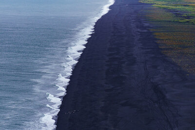 A sweeping view shows waves rolling in along Iceland’s Dyrhólaey Black Sand Beach, where a long, dark shoreline curves into the distance beside gray-blue ocean water, green coastal fields, and misty cliffs under an overcast sky.