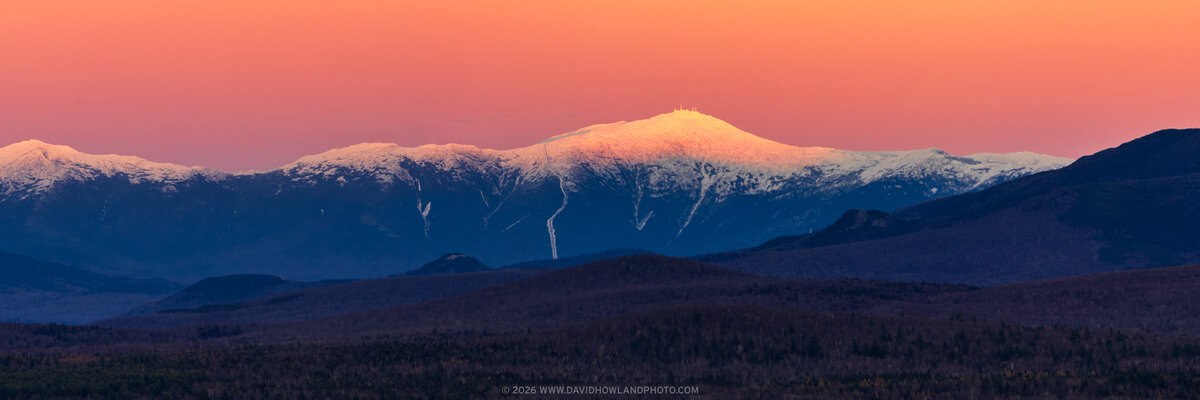 Mount Washington's snow-covered peaks are illuminated with warm orange and golden alpenglow against a peachy-orange sky at sunset or sunrise, with dark blue shadowed mountain ridges and forested foothills in the foreground.