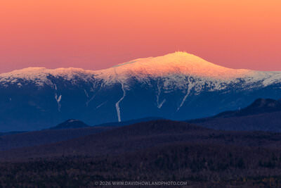 Mount Washington's snow-covered peaks are illuminated with warm orange and golden alpenglow against a peachy-orange sky at sunset or sunrise, with dark blue shadowed mountain ridges and forested foothills in the foreground.
