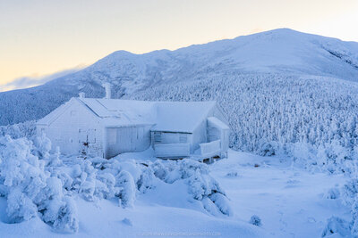 A snow-covered Greenleaf Hut with frost-laden evergreen trees in the foreground stands against a backdrop of the blue-tinted peak of Mount Lafayette during dawn.