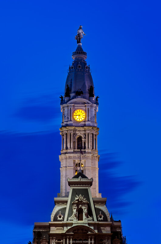 Philadelphia City Hall's ornate tower photographed at dusk, featuring an illuminated yellow clock face and the William Penn statue at its peak, with the building's detailed stonework and green mansard roof lit against a deep blue evening sky.