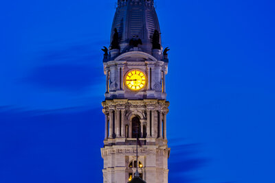 Philadelphia City Hall's ornate tower photographed at dusk, featuring an illuminated yellow clock face and the William Penn statue at its peak, with the building's detailed stonework and green mansard roof lit against a deep blue evening sky.