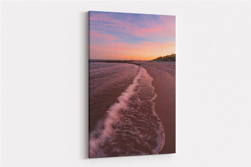 A gentle wave washes along a sandy beach shoreline at sunset in Chatham, MA, with warm pink and orange light reflecting off the water, a rocky jetty visible in the distance, and grass-covered dunes lining the right side beneath a dramatic pink and blue sky.