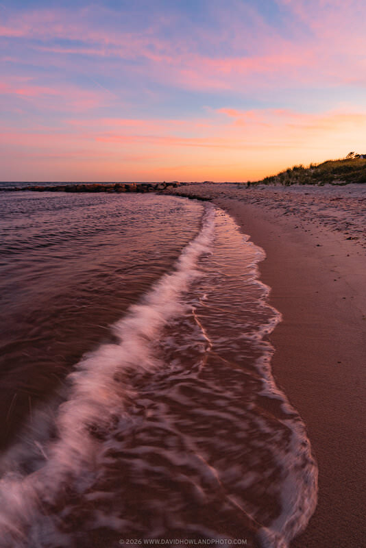 A gentle wave washes along a sandy beach shoreline at sunset in Chatham, MA, with warm pink and orange light reflecting off the water, a rocky jetty visible in the distance, and grass-covered dunes lining the right side beneath a dramatic pink and blue sky.
