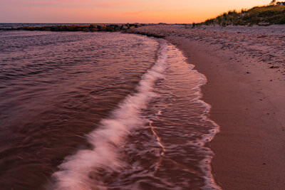 A gentle wave washes along a sandy beach shoreline at sunset in Chatham, MA, with warm pink and orange light reflecting off the water, a rocky jetty visible in the distance, and grass-covered dunes lining the right side beneath a dramatic pink and blue sky.