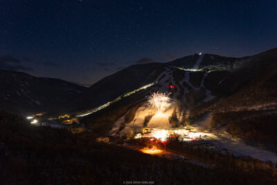 A photo of fireworks exploding over snow-covered Cannon Mountain under a clear winter sky filled with stars
