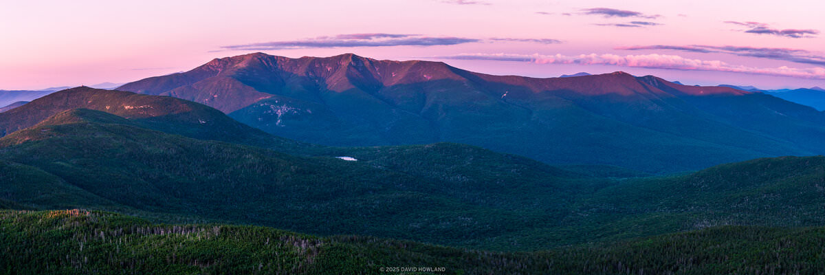 The image shows a panoramic view of mountain ranges at sunset, captured in New Hampshire's Franconia Notch region. The foreground features forested slopes covered in dense evergreen trees, while the dramatic peaks of the mountains rise against a pink and purple twilight sky. A small lake or pond is visible nestled in the valley between the mountains. The scene is captured during summer, with lush vegetation throughout. The composition emphasizes the scale and grandeur of the landscape through its wide format and layered perspective, from the foreground forest through multiple mountain ridges receding into the distance. The pink-hued sky creates a striking contrast with the deep blue-green tones of the mountains and forest below.