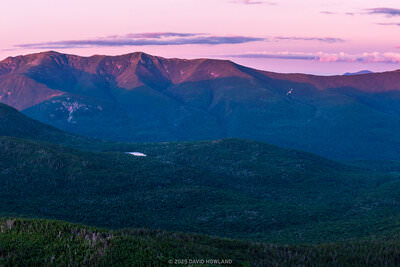 The image shows a panoramic view of mountain ranges at sunset, captured in New Hampshire's Franconia Notch region. The foreground features forested slopes covered in dense evergreen trees, while the dramatic peaks of the mountains rise against a pink and purple twilight sky. A small lake or pond is visible nestled in the valley between the mountains. The scene is captured during summer, with lush vegetation throughout. The composition emphasizes the scale and grandeur of the landscape through its wide format and layered perspective, from the foreground forest through multiple mountain ridges receding into the distance. The pink-hued sky creates a striking contrast with the deep blue-green tones of the mountains and forest below.