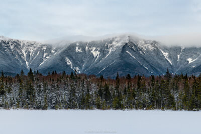 A panorama photo of the snow covered Franconia Ridge mountains above a pine forest and frozen Lonesome Lake in Franconia Notch, New Hampshire.