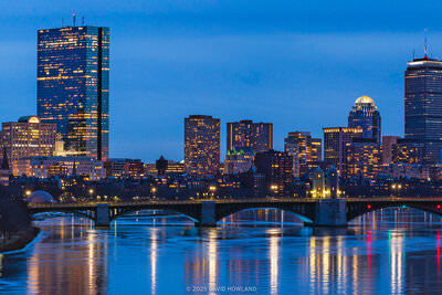 A panorama photo of the city lights of the Boston skyline and the Longfellow Bridge spanning a frozen Charles River at dusk