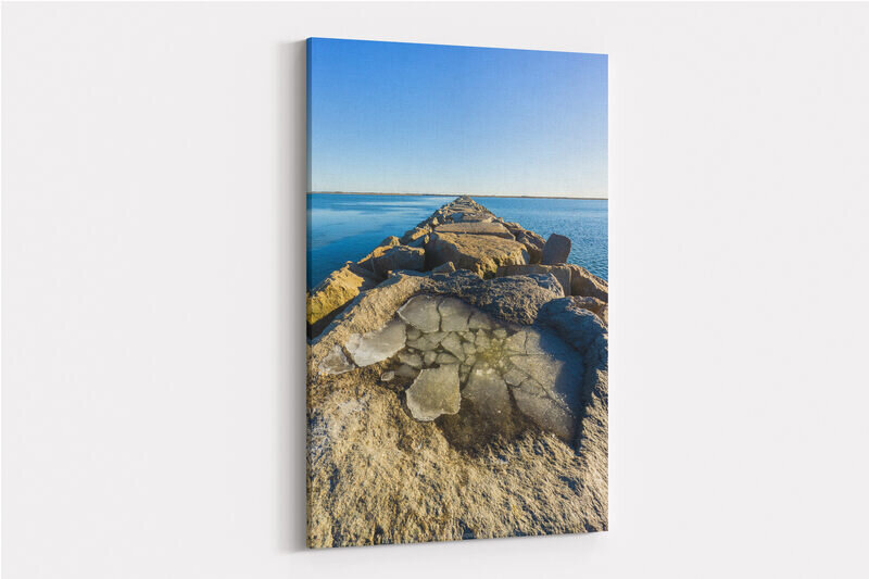 A rocky breakwater extends into calm blue waters of Provincetown Harbor on a cold winter day, with sheets of ice frozen in puddles between the weathered stone blocks and a clear blue sky above.