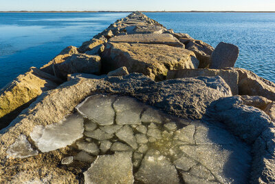 A rocky breakwater extends into calm blue waters of Provincetown Harbor on a cold winter day, with sheets of ice frozen in puddles between the weathered stone blocks and a clear blue sky above.