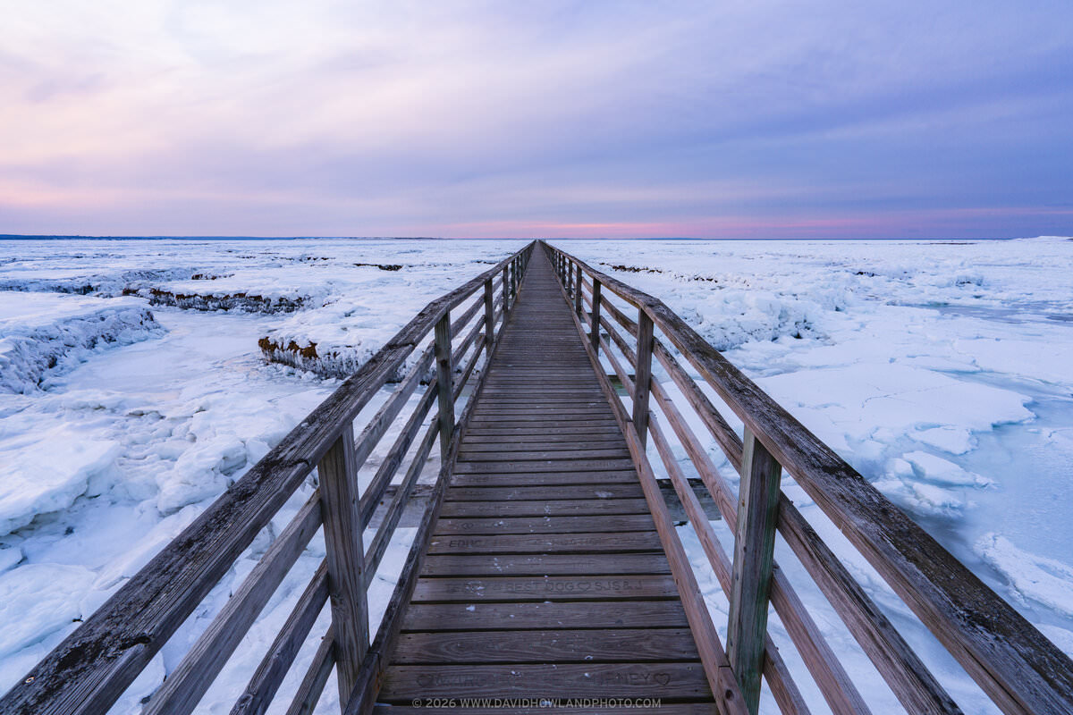A wooden boardwalk with railings extends toward the horizon through a frozen winter marsh landscape covered in snow and ice, beneath a pastel sunset sky transitioning from pink to blue.