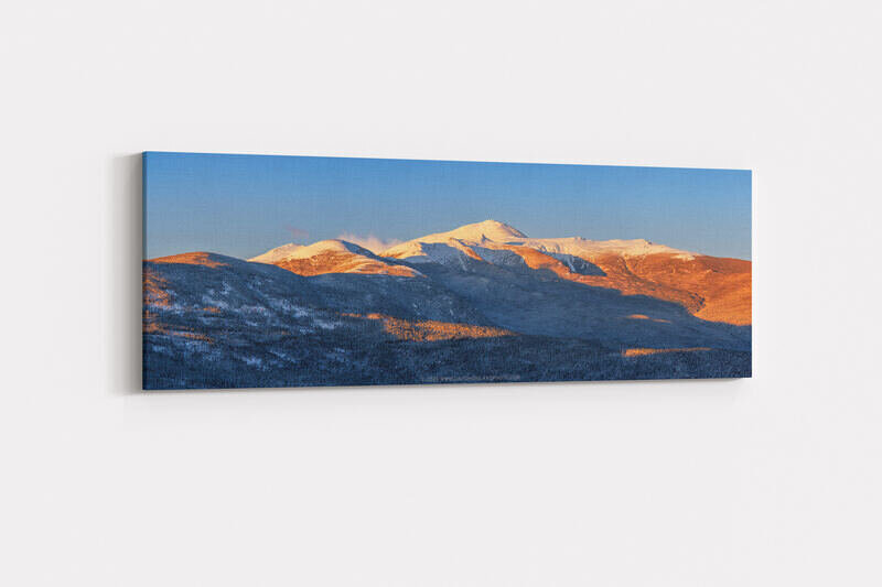 A panoramic golden hour view of New Hampshire's Presidential Range, with warm orange sunlight illuminating the snow-covered upper slopes and summits of Mounts Eisenhower, Pierce, Monroe, and Washington and the forested lower ridges, set against a clear deep blue winter sky.