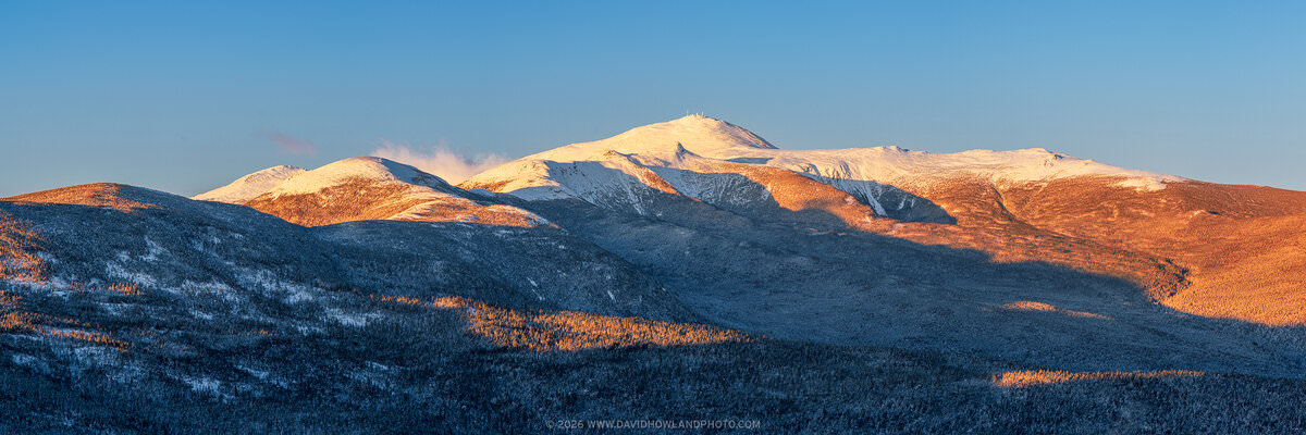A panoramic golden hour view of New Hampshire's Presidential Range, with warm orange sunlight illuminating the snow-covered upper slopes and summits of Mounts Eisenhower, Pierce, Monroe, and Washington and the forested lower ridges, set against a clear deep blue winter sky.