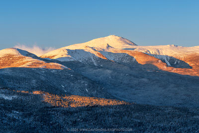 A panoramic golden hour view of New Hampshire's Presidential Range, with warm orange sunlight illuminating the snow-covered upper slopes and summits of Mounts Eisenhower, Pierce, Monroe, and Washington and the forested lower ridges, set against a clear deep blue winter sky.