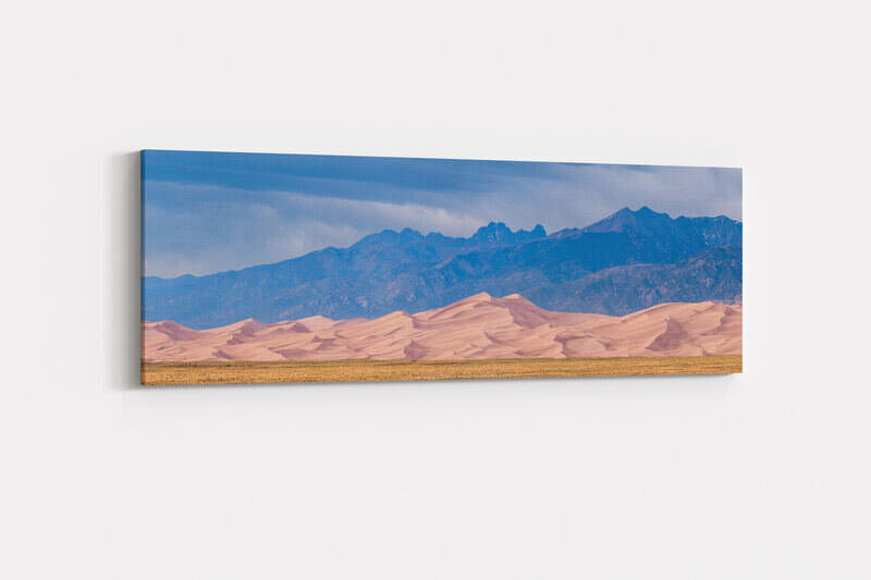 A wide panoramic landscape photograph of Great Sand Dunes National Park in Colorado, showing sweeping pink-tan sand dunes in the foreground rising against the dramatic, jagged Crestone Peaks of the Sangre de Cristo Mountains under a stormy blue-gray sky with visible rain in the distance.