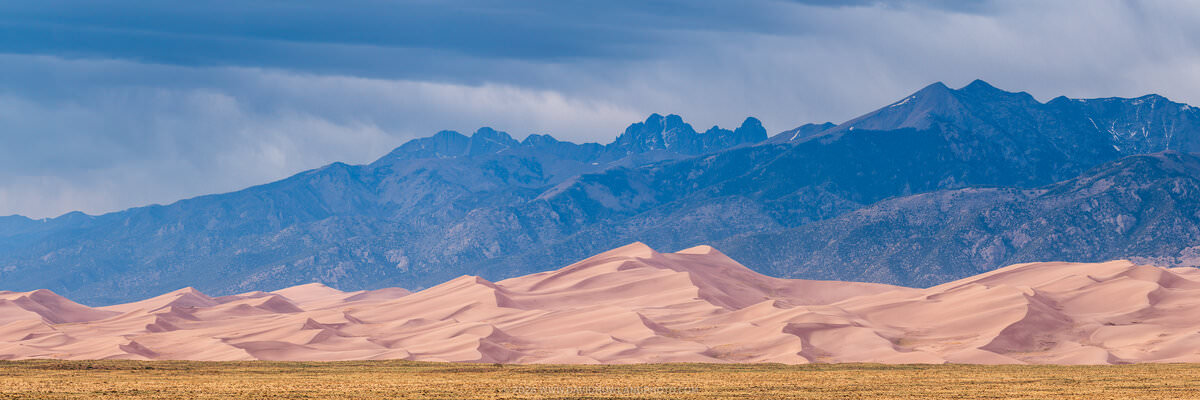 A wide panoramic landscape photograph of Great Sand Dunes National Park in Colorado, showing sweeping pink-tan sand dunes in the foreground rising against the dramatic, jagged Crestone Peaks of the Sangre de Cristo Mountains under a stormy blue-gray sky with visible rain in the distance.