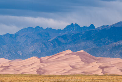 A wide panoramic landscape photograph of Great Sand Dunes National Park in Colorado, showing sweeping pink-tan sand dunes in the foreground rising against the dramatic, jagged Crestone Peaks of the Sangre de Cristo Mountains under a stormy blue-gray sky with visible rain in the distance.