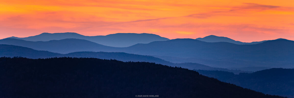 A colorful panorama photo of an orange sunset behind the layered blue-hued ridges of the Green Mountains of Vermont.