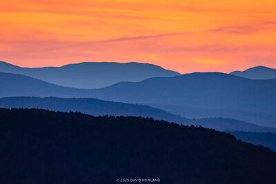 A colorful panorama photo of an orange sunset behind the layered blue-hued ridges of the Green Mountains of Vermont.
