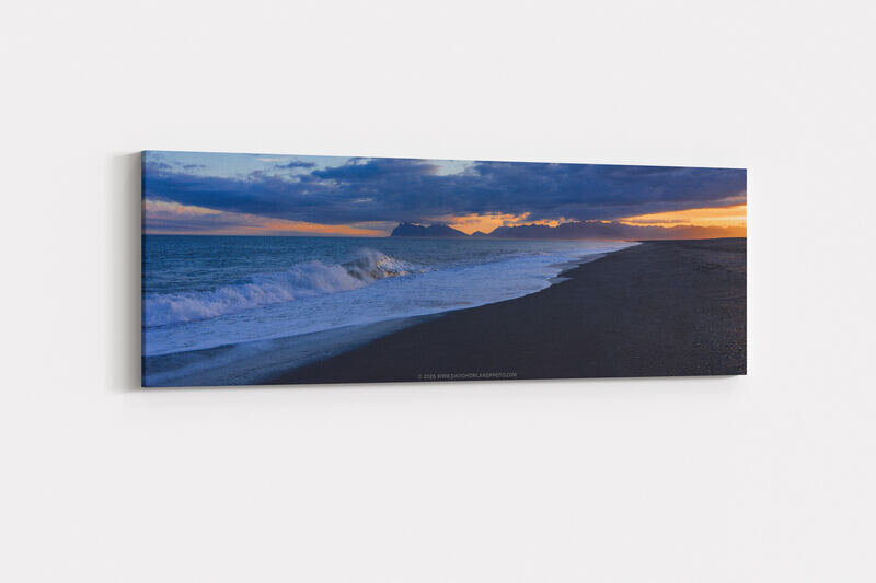 A dramatic sunset lights Hvalnes Beach in Iceland, where rough waves crash against dark sand in the foreground, with rugged coastline and distant mountains silhouetted beneath a glowing orange and pink sky.