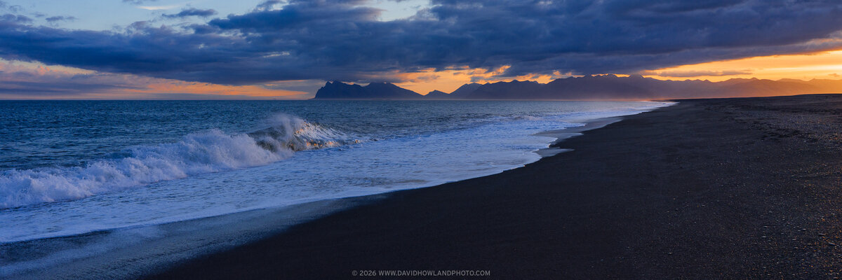 A dramatic sunset lights Hvalnes Beach in Iceland, where rough waves crash against dark sand in the foreground, with rugged coastline and distant mountains silhouetted beneath a glowing orange and pink sky.