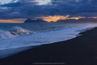 A dramatic sunset lights Hvalnes Beach in Iceland, where rough waves crash against dark sand in the foreground, with rugged coastline and distant mountains silhouetted beneath a glowing orange and pink sky.